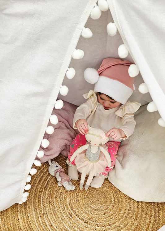 Child playing inside a white teepee with a pink hat and toy on a woven floor.